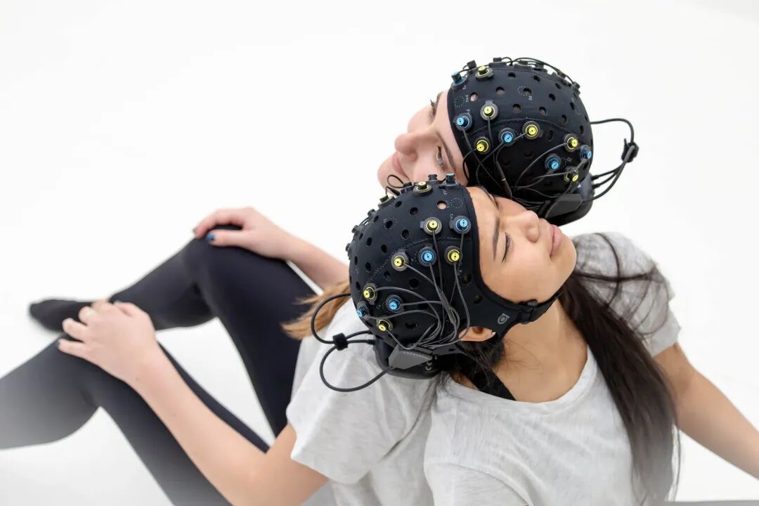 Two women wearing black fNIRS headcaps with multiple wires and sensors, sitting back-to-back with eyes closed on a white background.