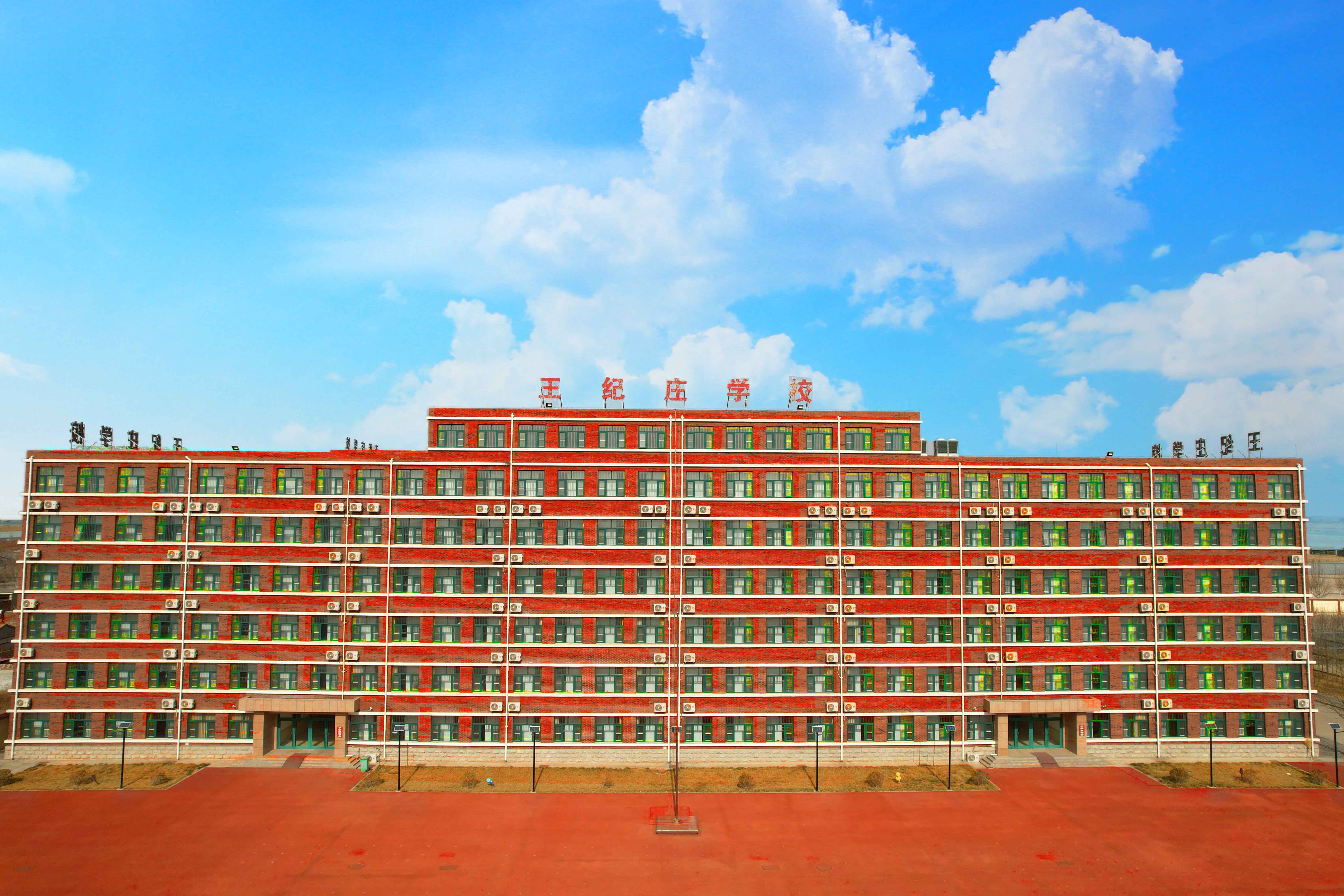 红砖外墙、多层结构的教学楼，楼顶醒目“王纪庄学校”字样，窗框为绿色，外立面整齐排列空调外机，前方为红色塑胶操场，整体布局规整，体现标准化校园建设。